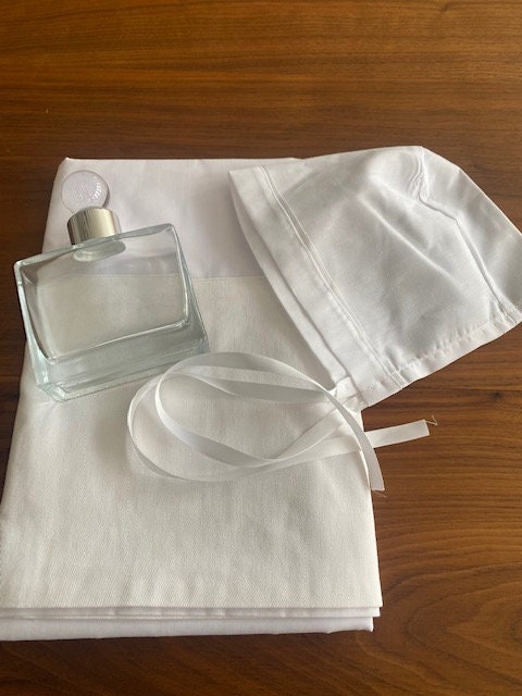 A white fabric bonnet with a white ribbon and a clear glass bottle with a white cap.. ladoset, oil cap, baptism towels, oil bottle, orthodox christening, christening towels, christening oil sheet , oil sheet, baptism oil sheet , Ladopana, personalised towels , oil sheet , bedding , Greek christening towel, Greek christening oil bottle ., christening oil cap