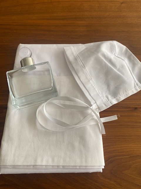 A white fabric bonnet with a white ribbon and a clear glass bottle with a white cap.. ladoset, oil cap, baptism towels, oil bottle, orthodox christening, christening towels, christening oil sheet , oil sheet, baptism oil sheet , Ladopana, personalised towels , oil sheet , bedding , Greek christening towel, Greek christening oil bottle .